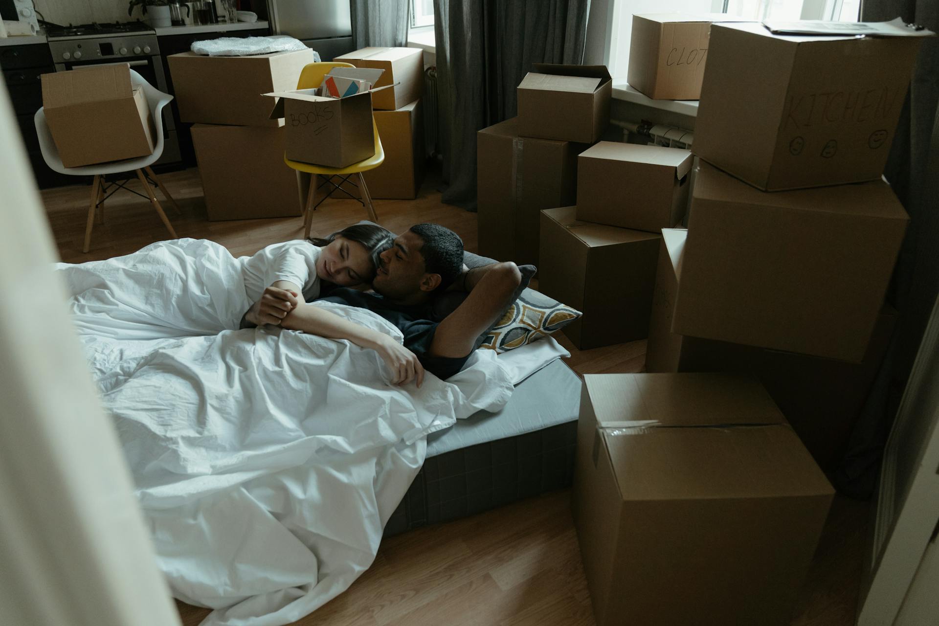 A couple lying on a mattress surrounded by moving boxes in their new home.