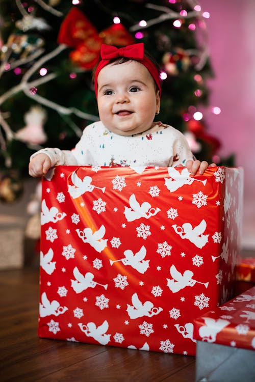 Adorable chunky baby girl with red bow on head sitting on floor behind wrapped gift box near decorated Christmas tree and smiling