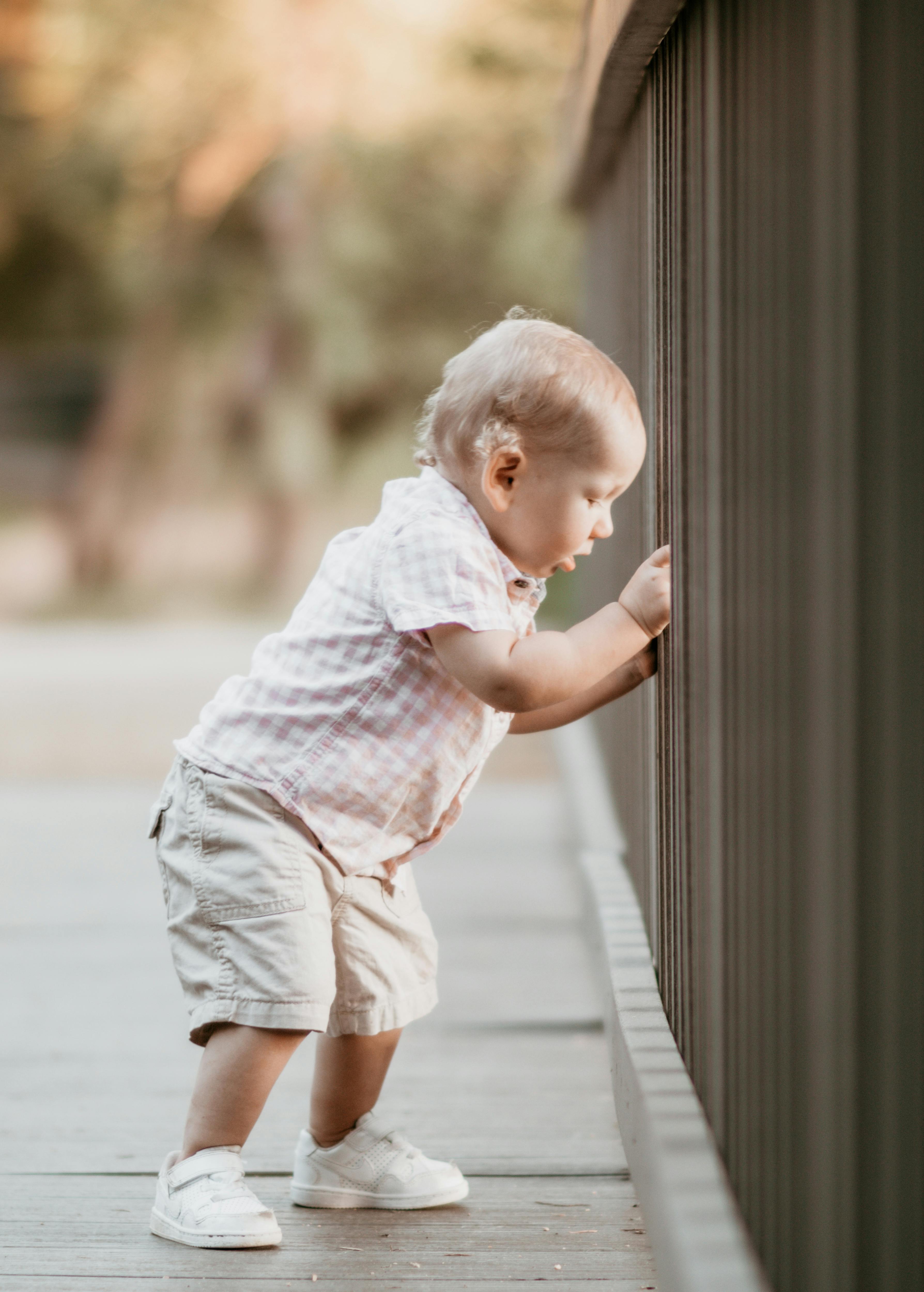 Side view of cute toddler in stylish outfit standing on wooden footbridge and looking down while playing in park