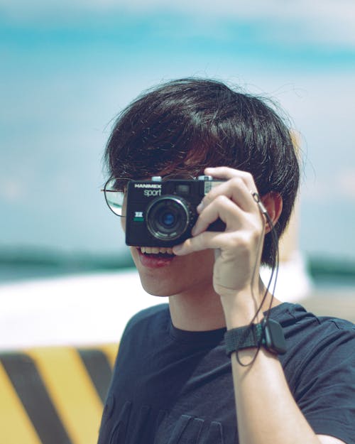 Unrecognizable young photographer in casual clothes and eyeglasses taking picture on vintage film camera on sunny day