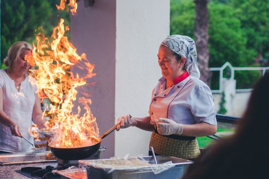 Happy middle aged female cook in uniform preparing delicious dish with flame in frying pan during work in outdoor restaurant located in green park