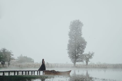 Free A woman in a black dress stands on a foggy dock beside a boat amidst serene nature. Stock Photo