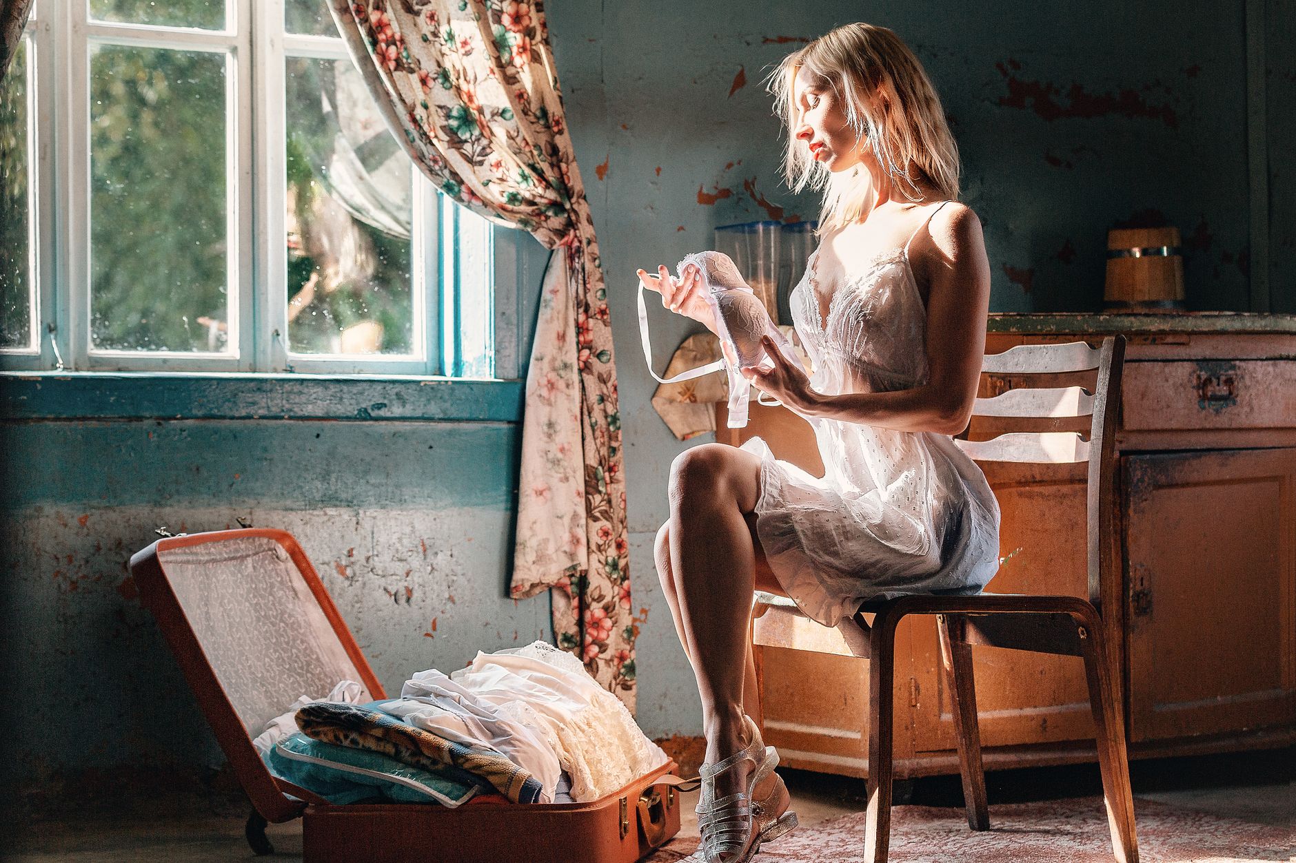 Side view of sensual young female in lace nightgown sitting on chair in old shabby house with bra in hands while packing suitcase