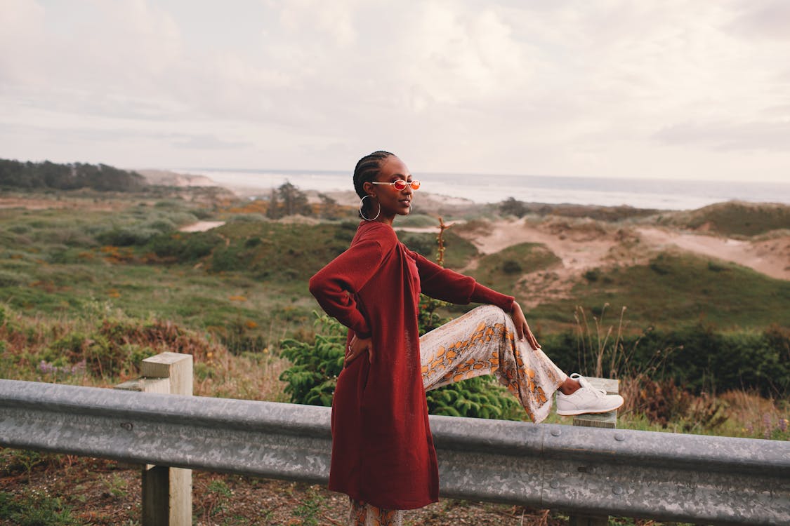 Free Side view of young stylish African American female traveler in trendy outfit and sunglasses sitting on road metal fence during trip near ocean against cloudy sky Stock Photo