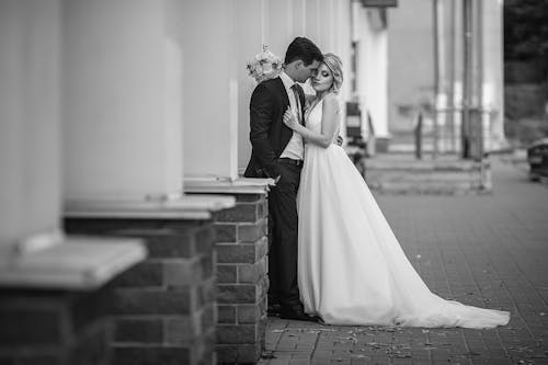 Free Black and white full body of unemotional bride and groom in elegant wedding suits embracing while standing on pavement next to row of white columns Stock Photo