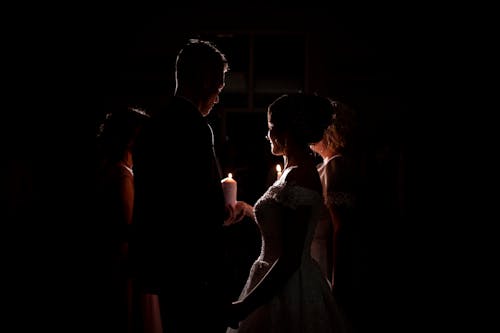 Free Side view of newlywed couple in wedding gown holding small white flaming candle while standing in dark room among group of people Stock Photo