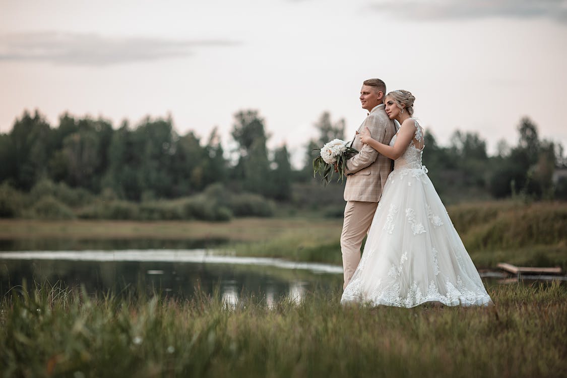 Full body of bride and groom hugging each other while standing on green meadow near lake in countryside in summer