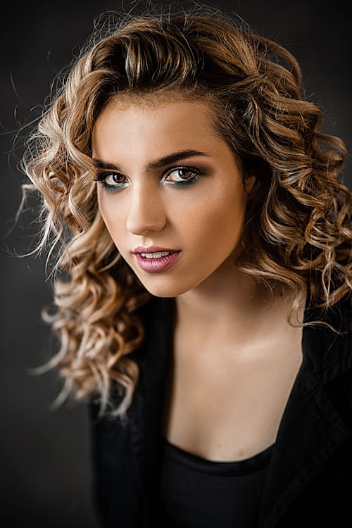 Free Attractive female with blond hair and bright makeup wearing black outfit and looking at camera against dark background in studio Stock Photo