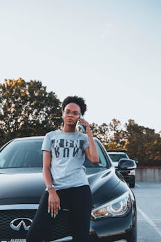 African American woman confidently leans on her car as she talks on the phone in a parking lot.