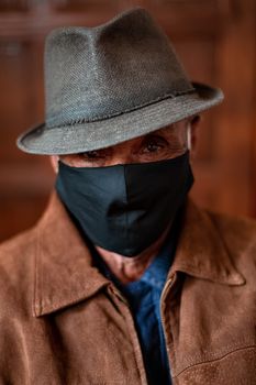 一位戴着时尚帽子和防护口罩的严肃年长男性，站在房间内模糊的木制背景前，注视着镜头