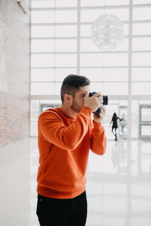 Free Side view of anonymous bearded male with orange sweater taking photo on camera while standing in spacious light studio in daylight Stock Photo