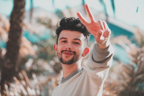 Cheerful male with black hair smiling and looking at camera while standing on blurred background and doing peace sign