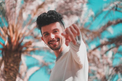 Low angle of modern trendy man with earring pointing with finger at camera standing under palm trees in sunshine