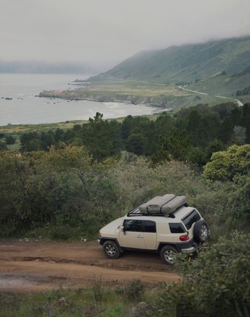 Free SUV car parked on dirty road near green trees and rocky shore of ocean against cloudy sky Stock Photo