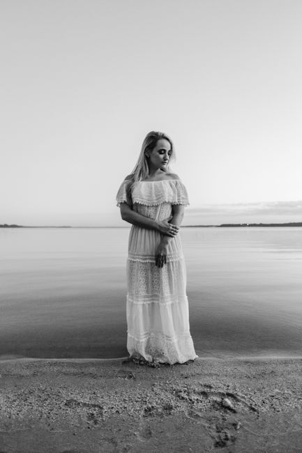 Full body of melancholic lady in long white dress standing peacefully on sandy beach looking down