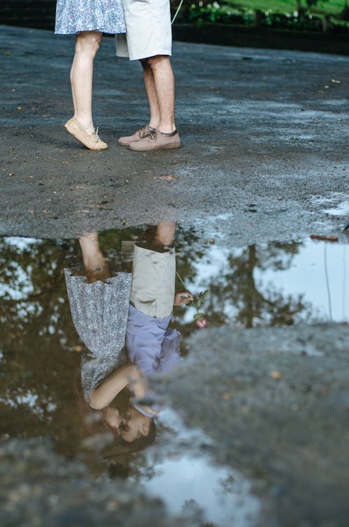 Full body of crop happy man and woman kissing and reflecting in puddle on asphalt road