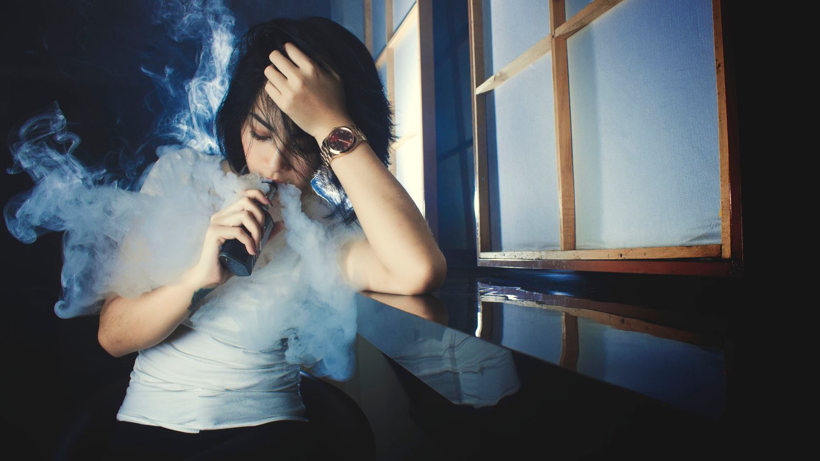 Cool casual brunette in white t shirt smoking electronic cigarette while leaning on hand in dark studio