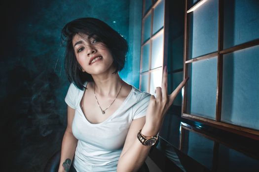 Young casual brunette with short hairstyle and in white t shirt showing two fingers and biting lips provocatively in dark room
