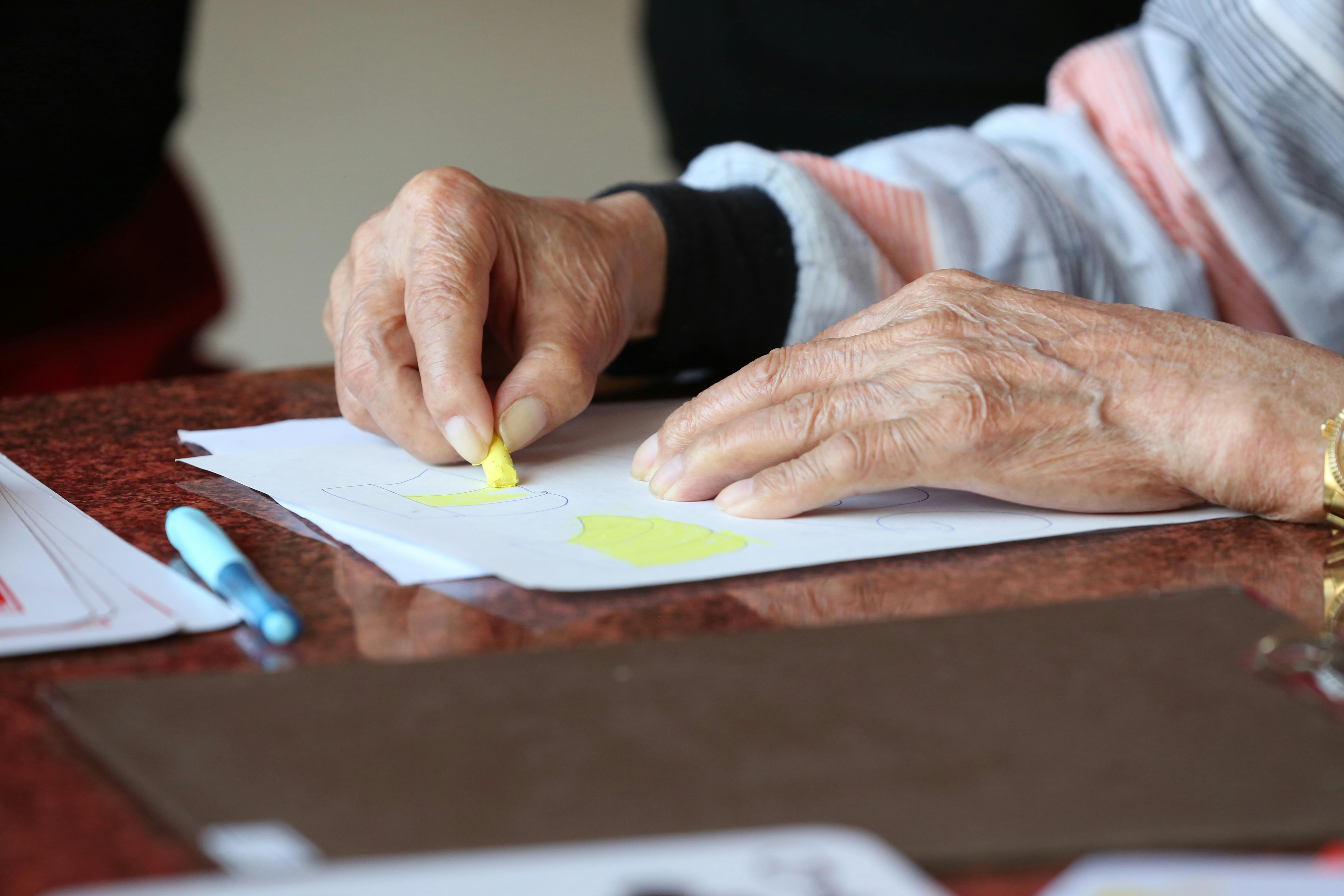 Faceless senior patient with arthritis drawing with yellow crayon at table in rest home