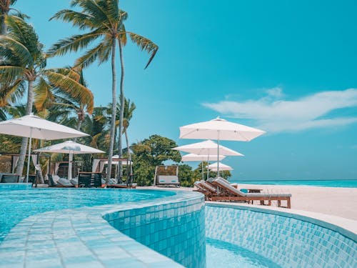 Tiled pool of exotic resort placed on white sandy beach with parasols and green palms