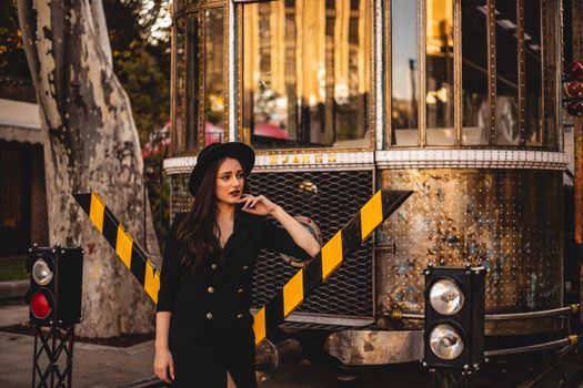 Stylish woman in a black outfit posing near a vintage tram in an urban setting. Fashion forward scene.
