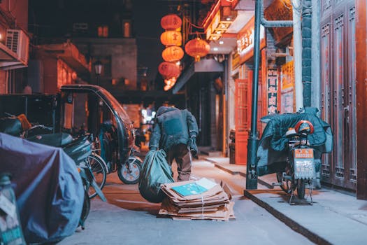 A person collecting cardboards in a vibrant Beijing alley at night, illuminated by red lanterns.