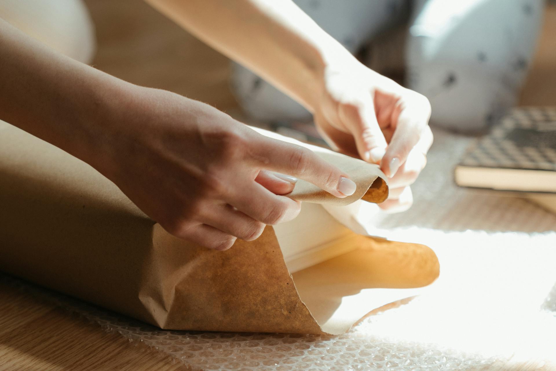 Hands wrapping an item in brown paper with sunlight streaming in, highlighting the texture and details.