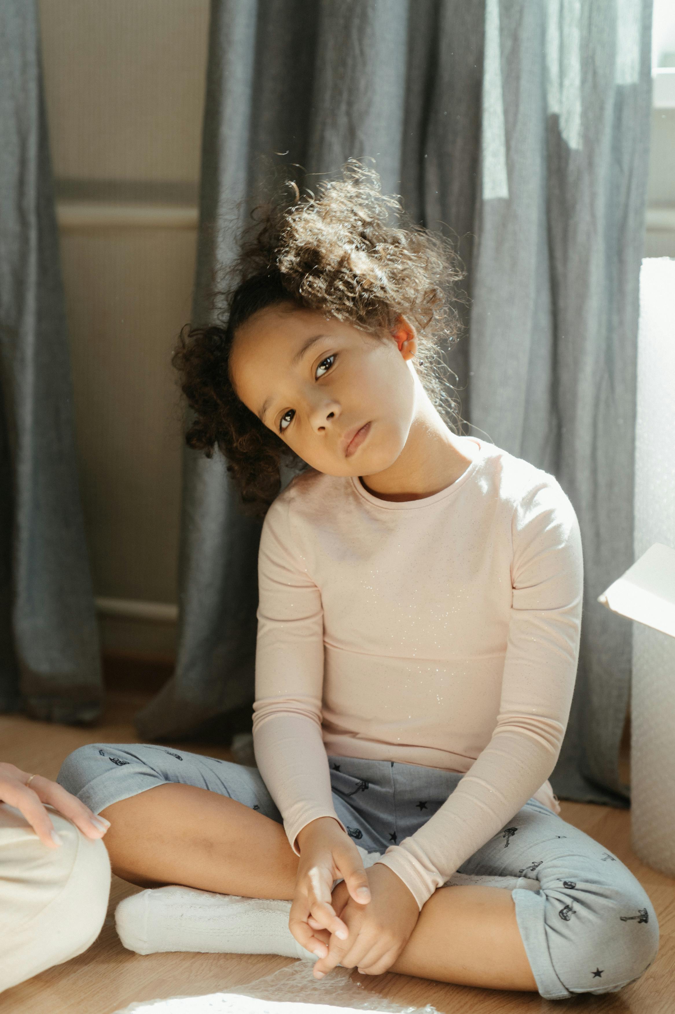 Young girl with curly hair sitting indoors in soft light, pondering deeply.