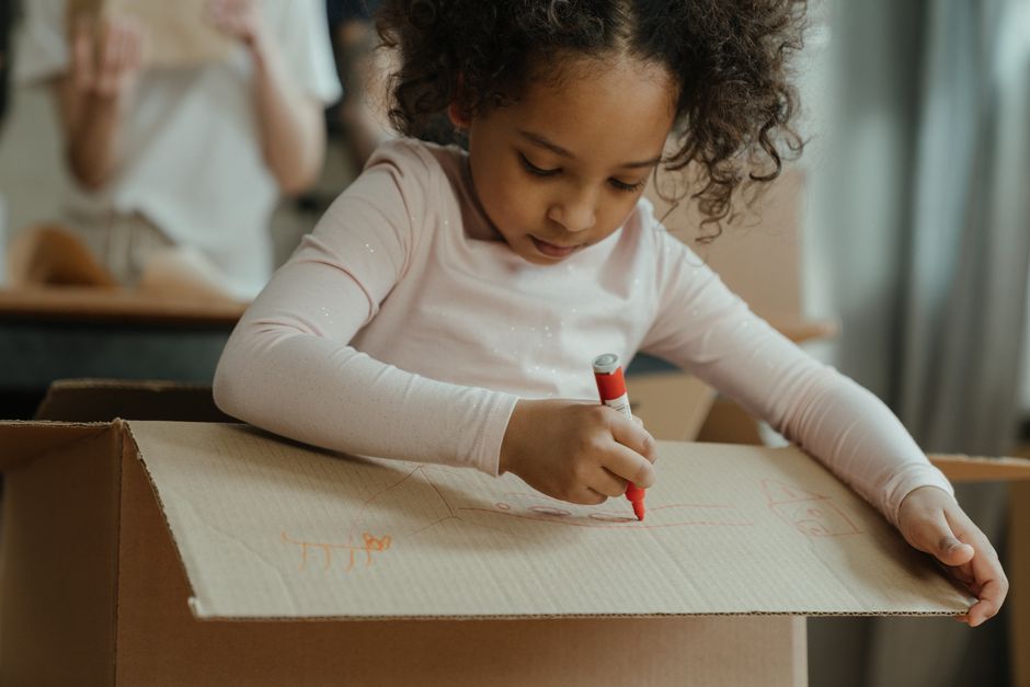 Young girl engrossed in drawing on a cardboard box during a creative indoor activity.