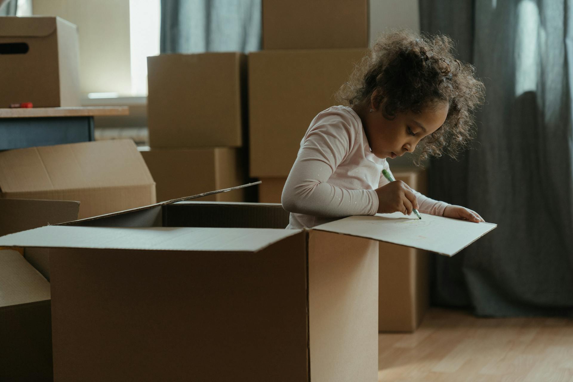 A young child creatively drawing while sitting in a cardboard box surrounded by moving boxes indoors.