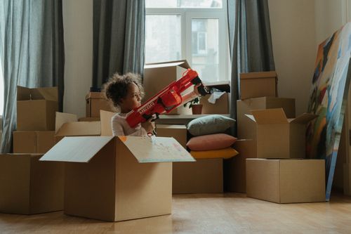 Free A child enjoying playtime with a toy gun amidst moving boxes, indoors. Stock Photo