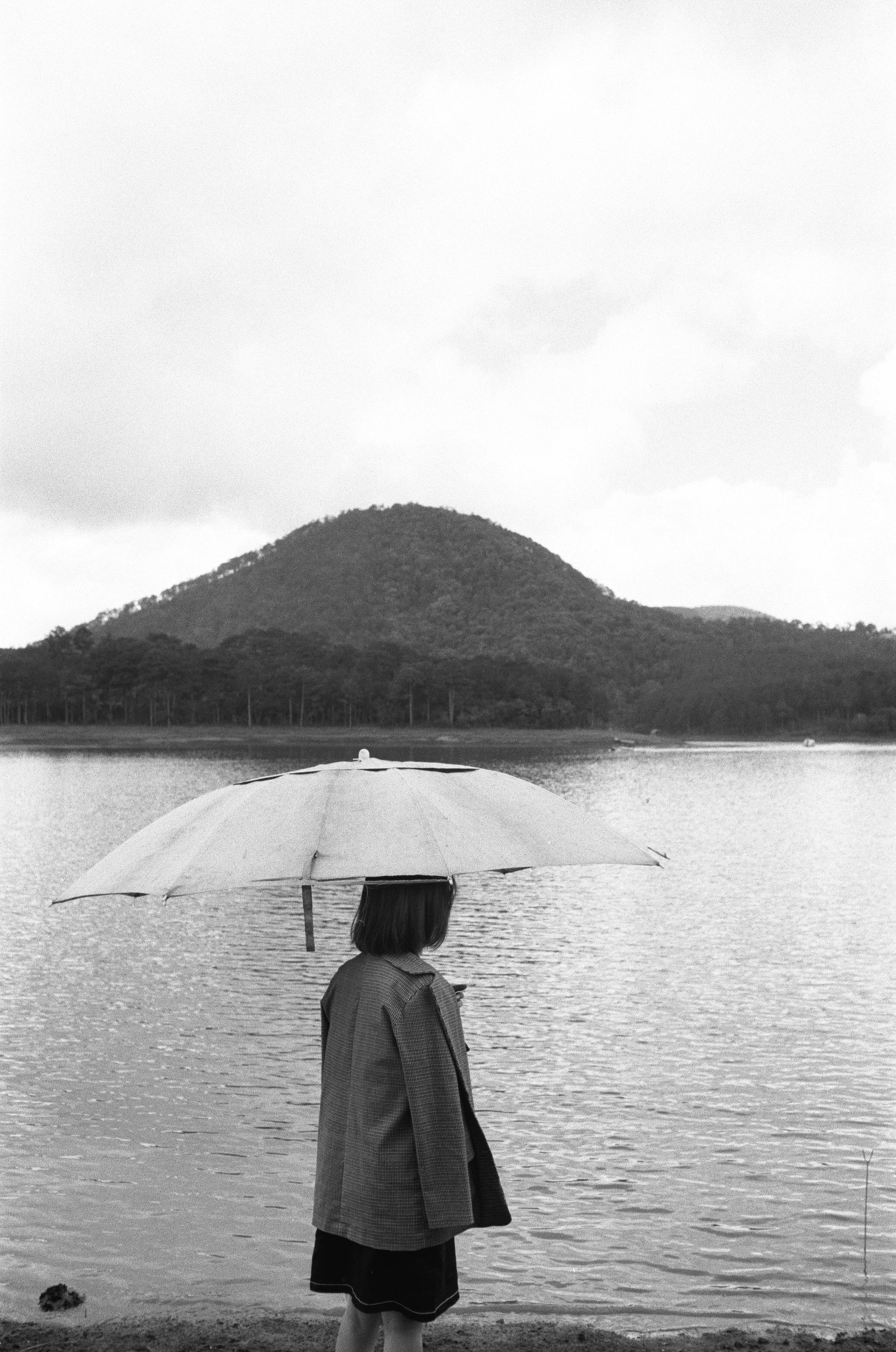 A woman stands with an umbrella near a calm lake surrounded by mountains in a grayscale scene.