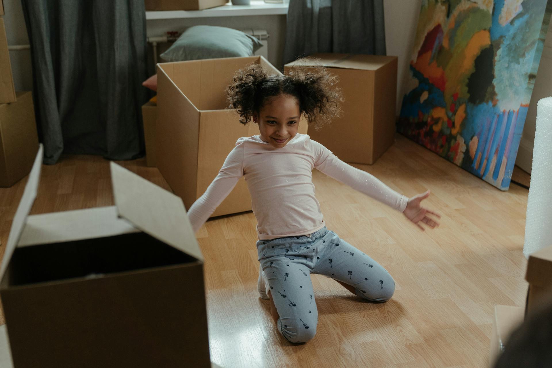 A happy child with curly hair plays with cardboard boxes in a new home. The scene depicts joy and exploration.