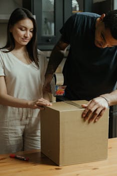 A couple prepares for moving day by sealing a cardboard box in their kitchen.