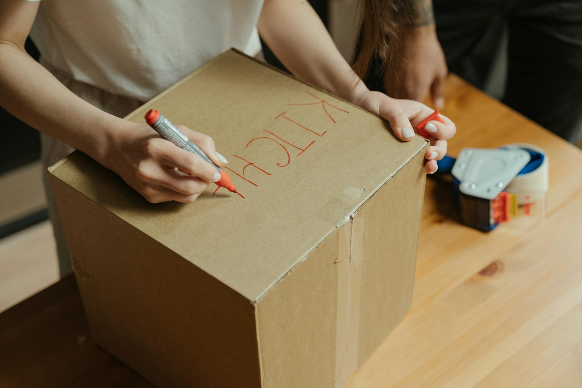 Person labeling a cardboard box 'Kitchen' while moving into a new home.