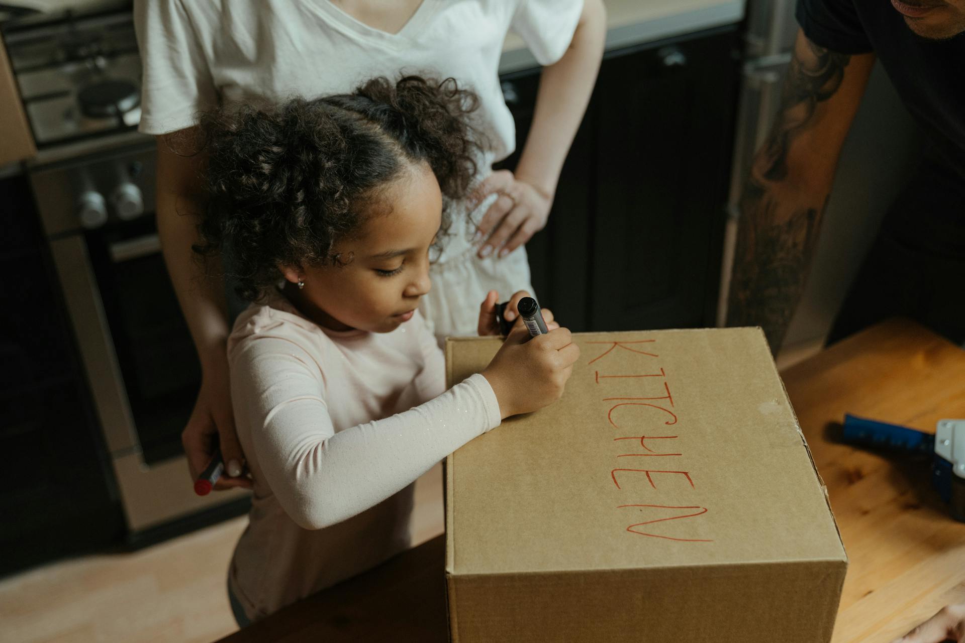 A mother and daughter label a cardboard box in their new kitchen, capturing the moving experience.