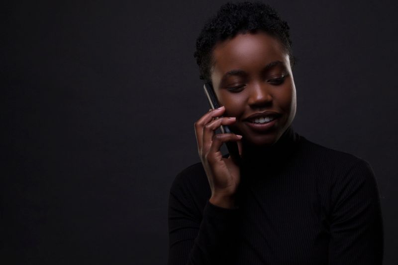 A Black woman with short natural hair holds a phone to her ear against a dark background, eyes lowered, listening with a slight smile.