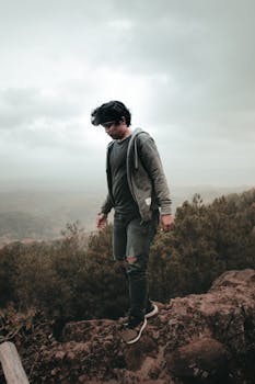 A young man in casual attire stands on a rocky cliff with a misty backdrop.