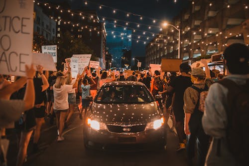 Modern car with glowing headlights riding on street amidst crowd of faceless people walking together during protest at night