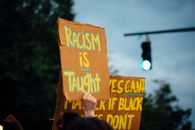 Crop anonymous activist demonstrating placard with racism is taught text while protesting for Black Lives Matter movement in evening