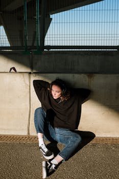 Woman with curly hair sitting against a wall in an urban environment, wearing casual attire.