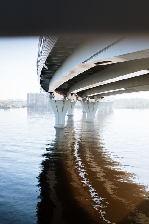 Free A sleek modern bridge spanning over a still river reflecting the urban environment on a clear day. Stock Photo