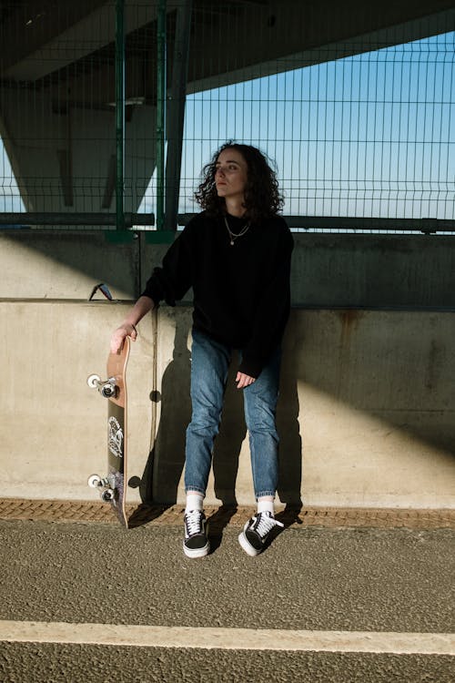 Woman in Black Long Sleeve Shirt and Blue Denim Jeans Sitting on Brown Wooden Skateboard