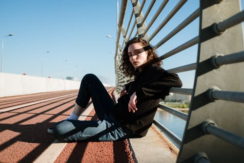 Free Girl sitting on an urban bridge, casting shadows in sunlight, reflecting a modern city vibe. Stock Photo