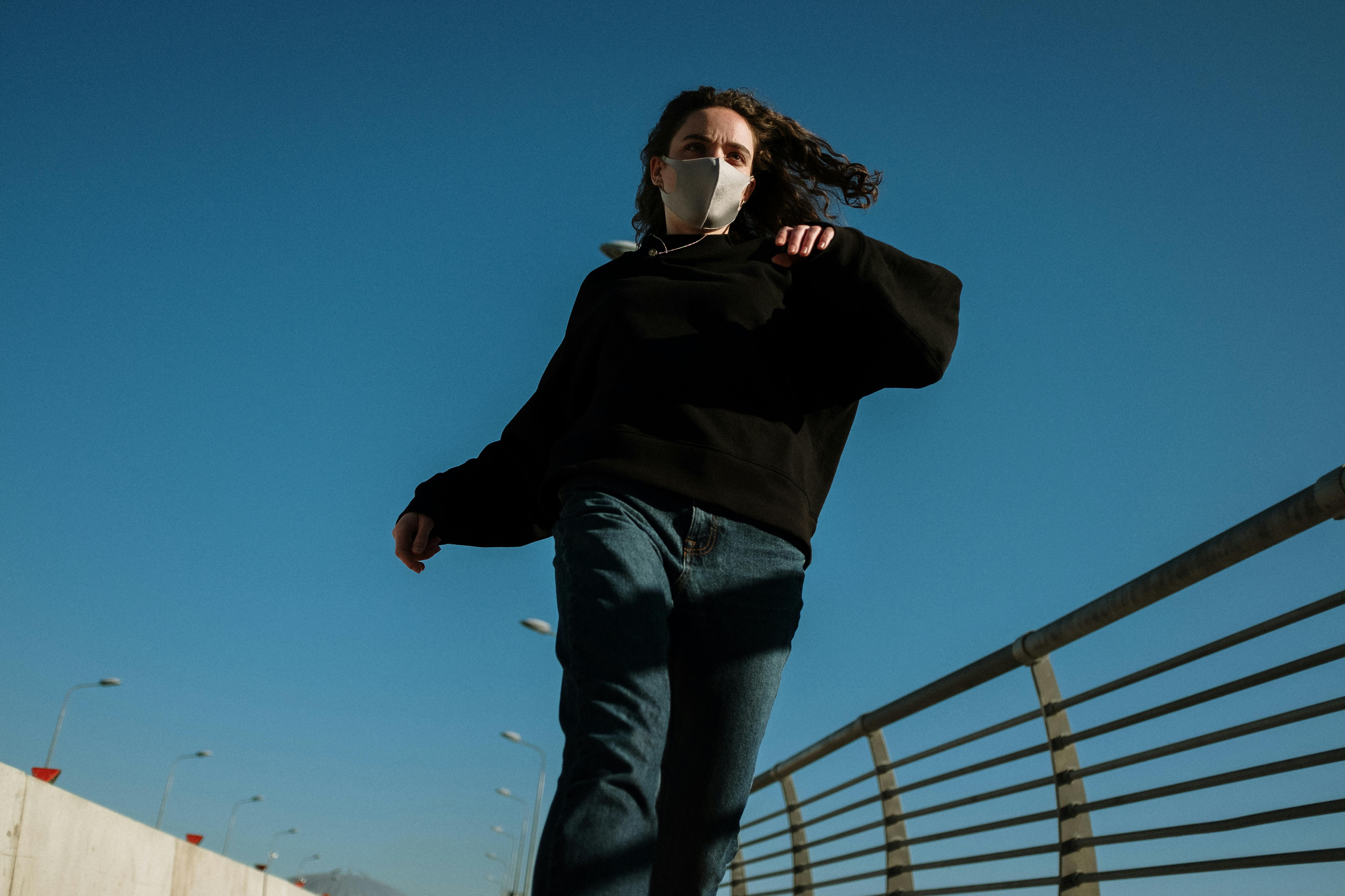 在晴朗的蓝天下，一个戴着口罩的人沿着城市桥梁奔跑，展现了现代城市生活