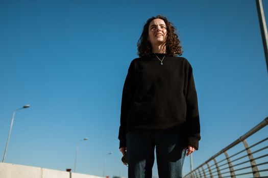 一位自信的年轻女子，卷发站在城市桥上，天空晴朗湛蓝