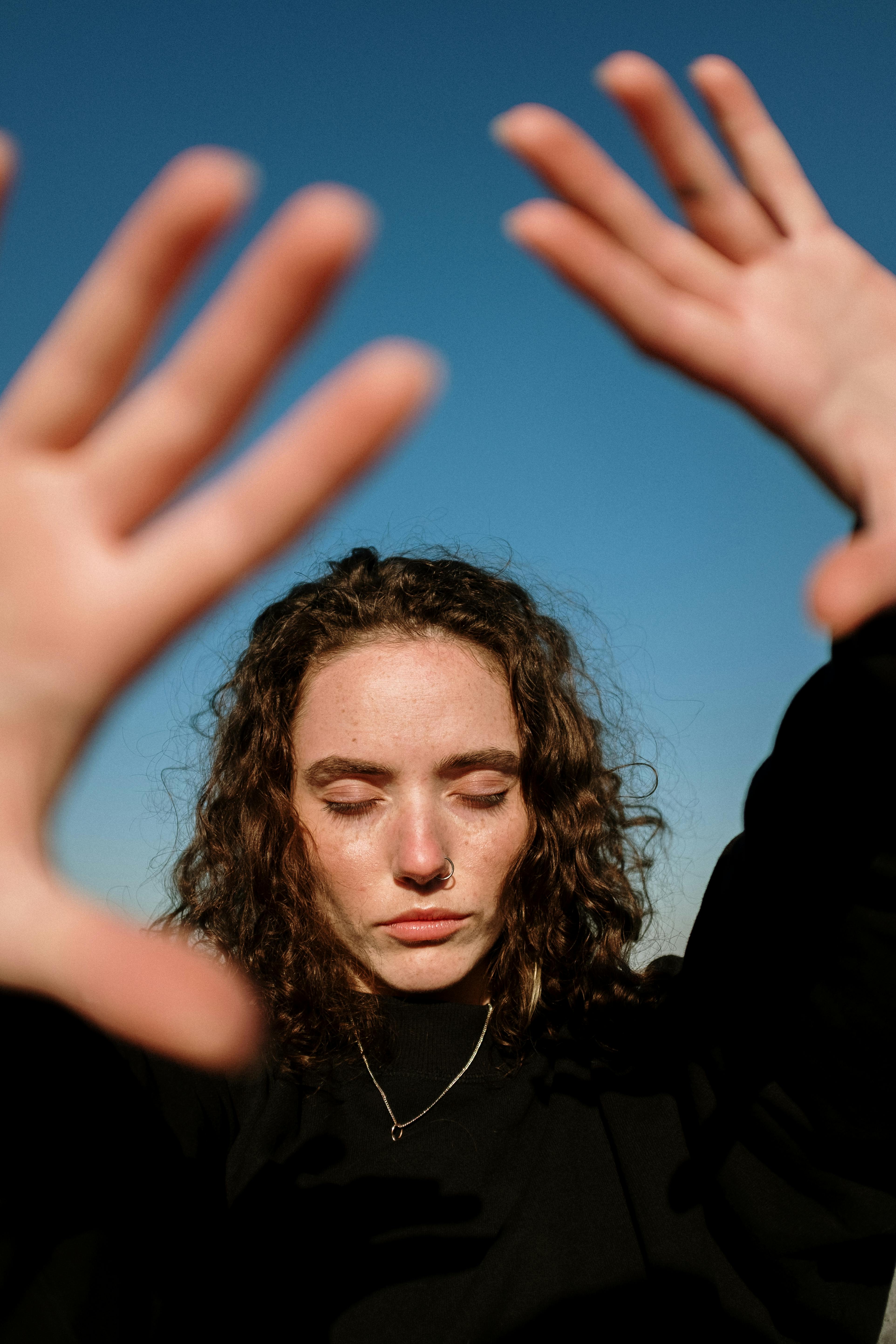 Portrait of a young woman with curly hair and nose piercing, hands raised, eyes closed under the blue sky.