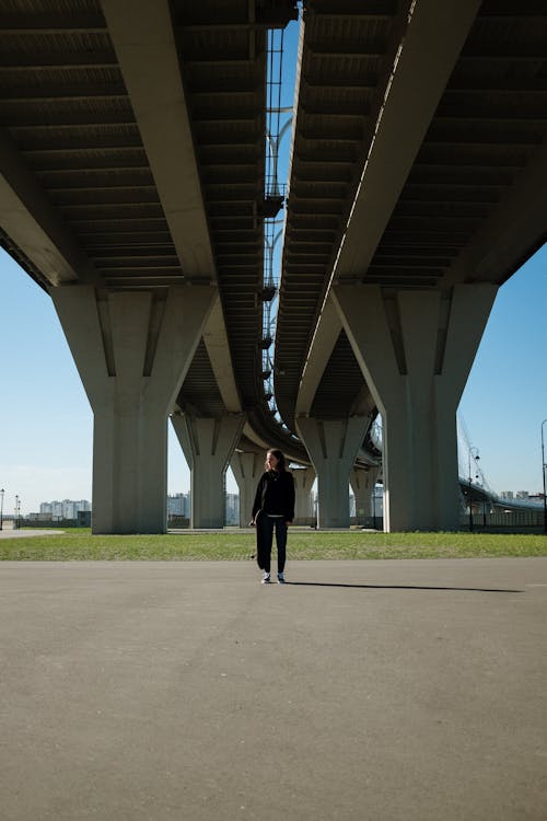 Person in Black Jacket and Black Pants Walking Under White Bridge