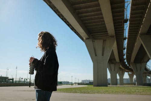 Woman in Black Jacket and Blue Denim Jeans Standing Under Bridge