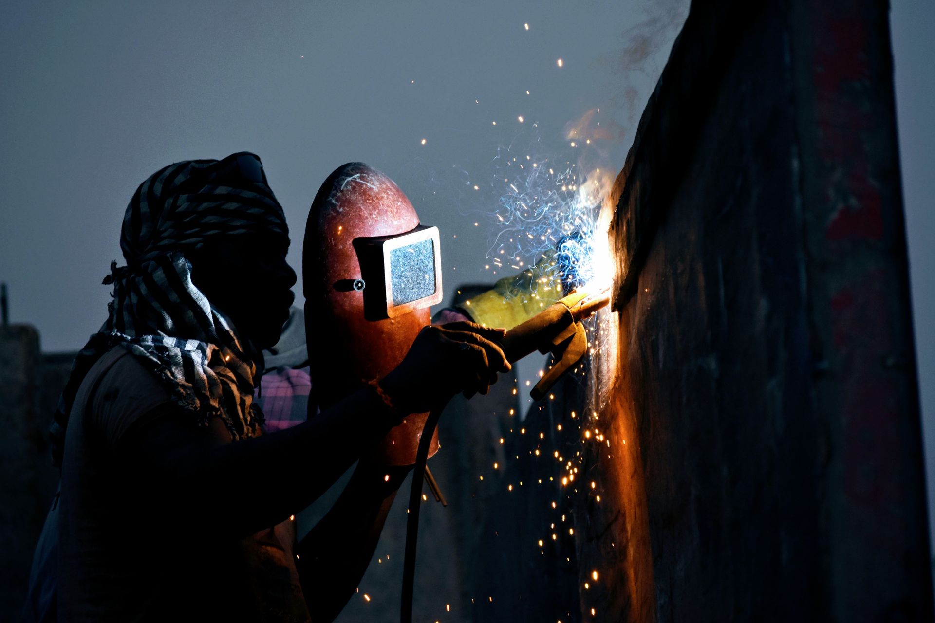 A silhouette of a welder working outdoors at dusk with sparks flying.